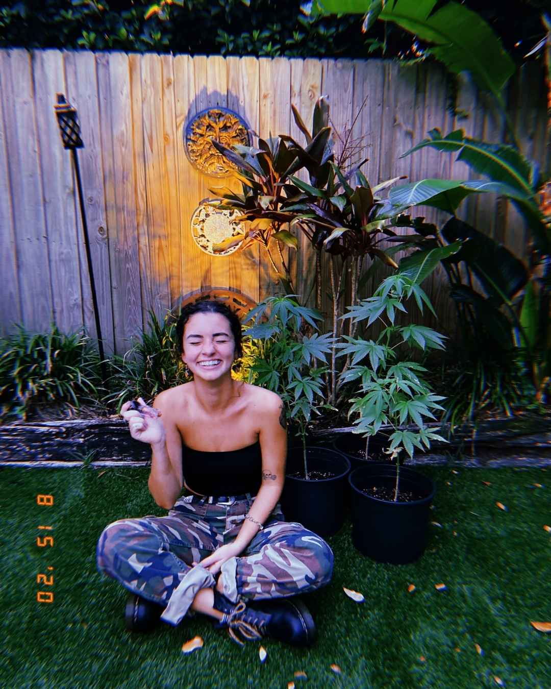 Smiling cannabis coach Isabella DeChard seated cross-legged in front of potted cannabis plants, holding a joint.