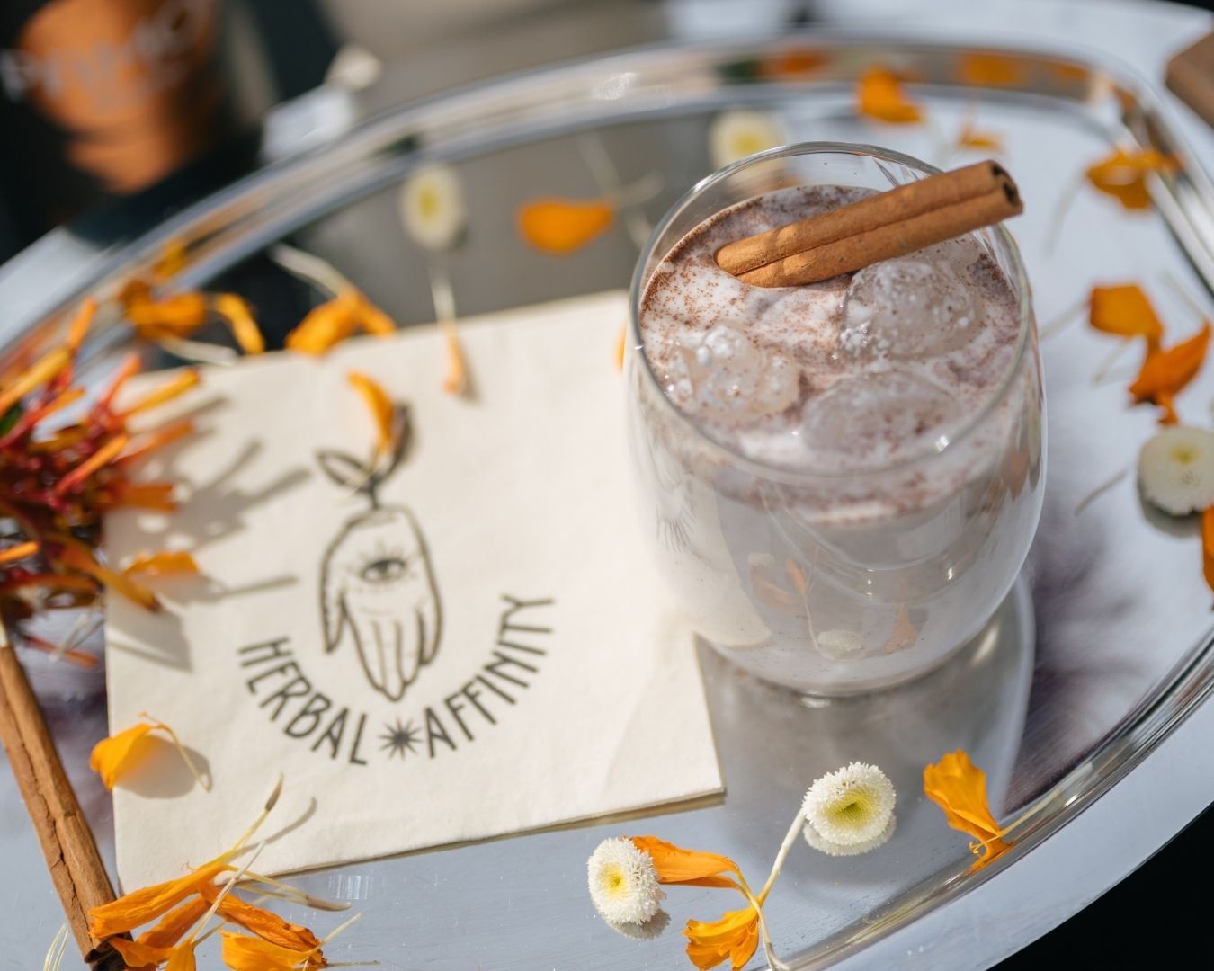 Cannabis beverage tasting setup featuring an iced cinnamon cannabis drink on a silver tray with Herbal Affinity branding and floral accents.