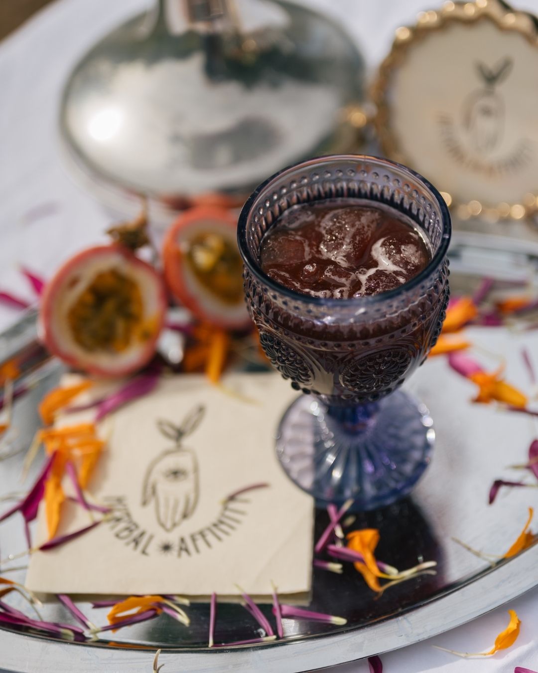 Decorative glass filled with a dark herbal mocktail over ice on a mirrored tray with passion fruit and scattered flower petals in natural light.